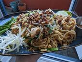 Pad Thai on a rectangular blue plate. There are bean sprouts and garlic chives to the left, a lime wedge and a cup of chili sugar to the right, and peanuts and fried shallots on top.